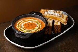 A close-up of creamy tomato soup drizzled with oil and cream swirls, served in a black pot alongside a grilled cheese sandwich cut into strips.