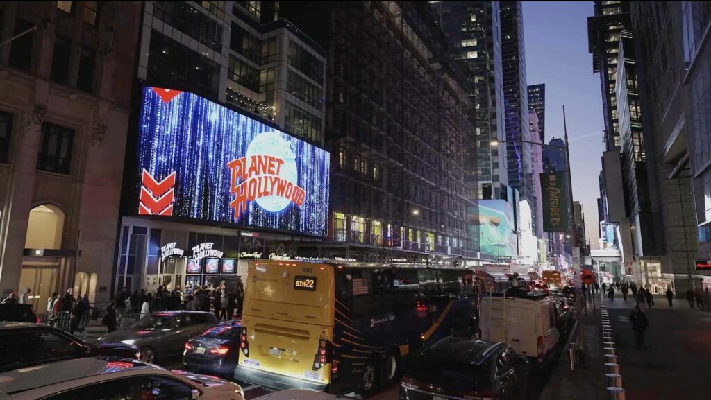 A large billboard in Times Square at night displays the glittering "Planet Hollywood" logo over a busy street filled with traffic and pedestrians.