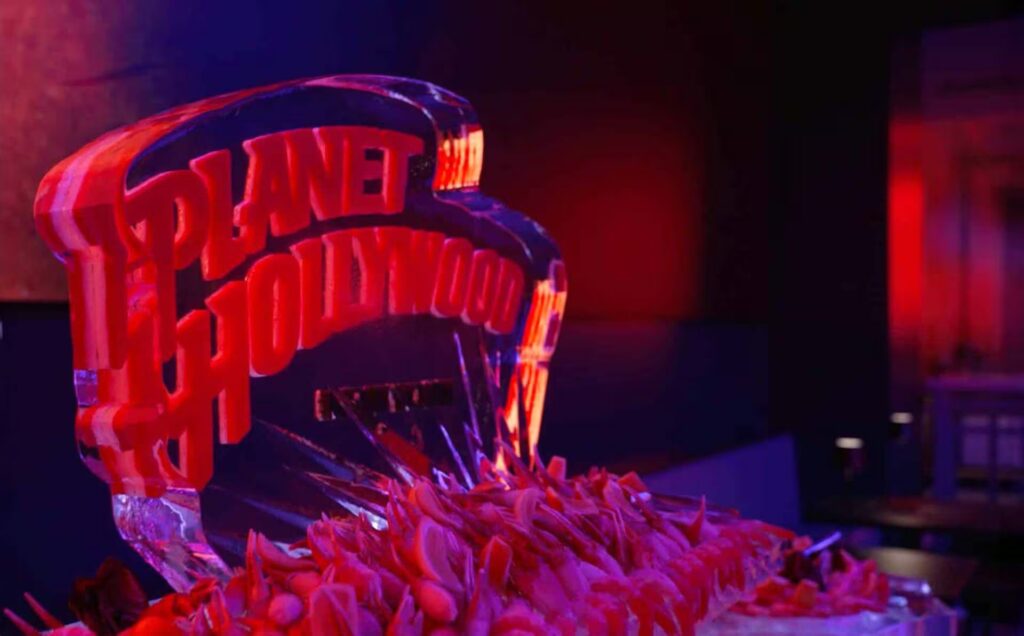 A close-up view of the Planet Hollywood New York ice sculpture, bathed in bright red light, with shrimp piled at its base.