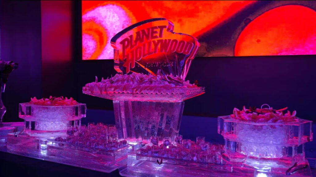 A shrimp cocktail display features a central Planet Hollywood New York ice sculpture, set against a red glowing screen.