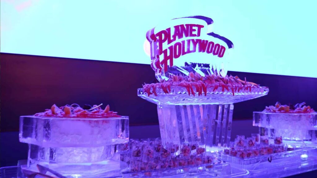 A grand ice sculpture centerpiece featuring the Planet Hollywood logo is surrounded by shrimp cocktails on ice platters, set against a bright blue screen.