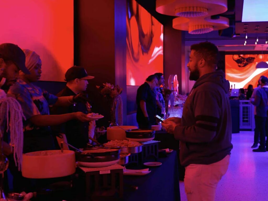 Staff serve a guest from a buffet line in a PHNYC event space illuminated by dramatic red and blue lighting from large wall screens.