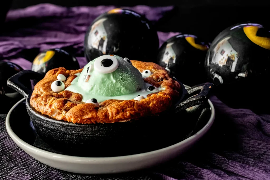 Monster Cookie Skillet topped with green ice cream and candy eyeballs in a spooky Halloween setting.