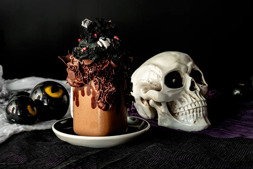 Midnight Chocolate Shake topped with chocolate frosting, sprinkles and spooky Halloween decorations beside a skull.