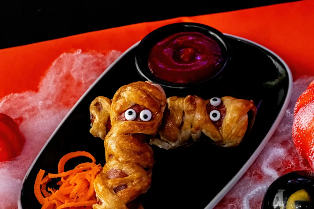 Kids Mummy Dog wrapped in golden dough with candy eyes, served with dipping sauce and a spooky Halloween presentation.